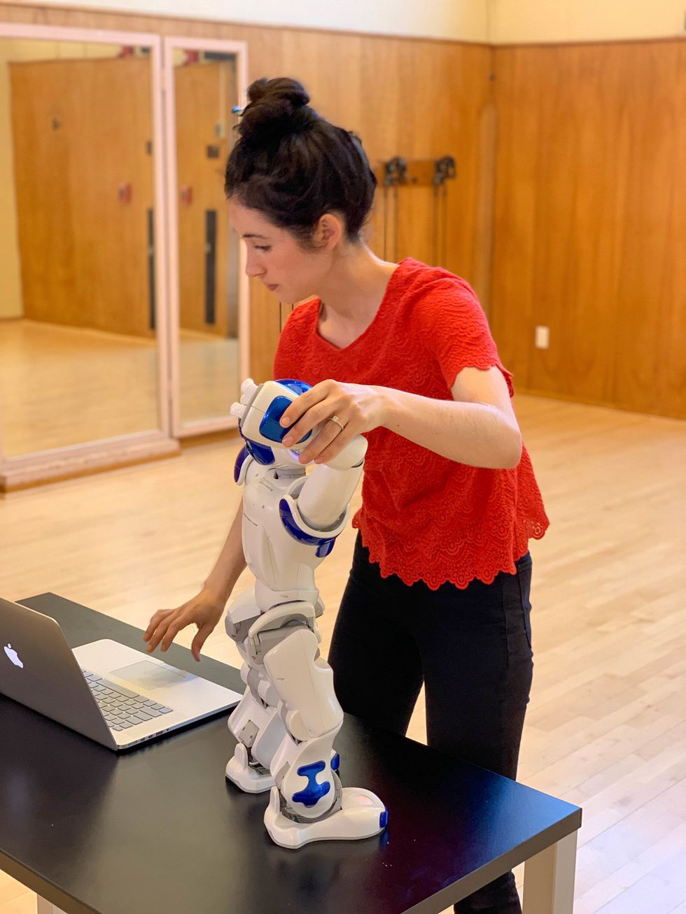 Catie Cuan holds a small robot standing on a table, while she types on a laptop.