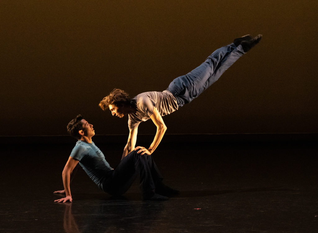 One dancer sits facing the side, hands pressed into the ground behind him for support as a second dancer is suspended almost parallel to the floor with one hand pressing against the first's knees. In this moment of suspended motion, the dancers stare intently into each other's eyes.