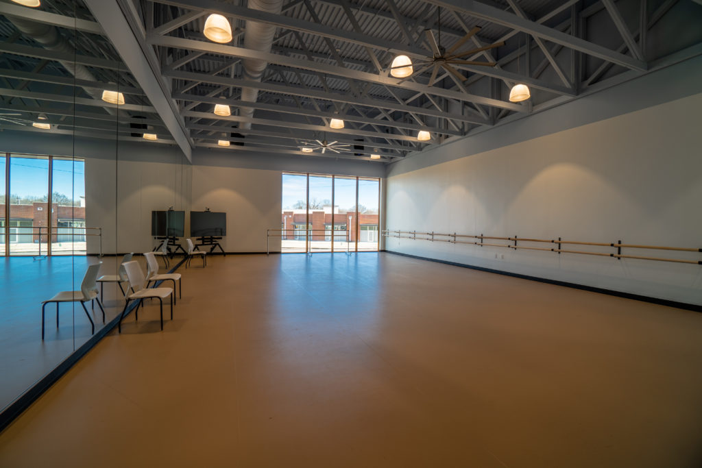 A large dance studio with a wall of mirrors, a wall of barres and tan-shaded floors