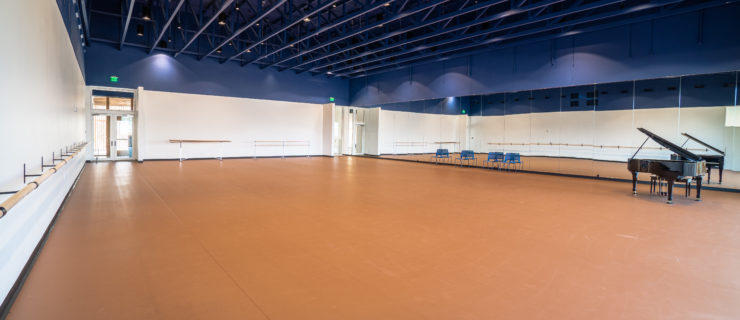 An extra large dance studio with blue beams in the ceiling, wall barres, a wall of mirrors and a piano