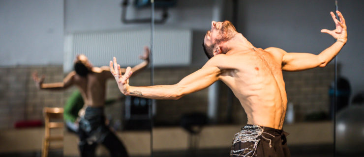 A shirtless male dancer sits into one hip, head arched back to look at the ceiling, his arms curving as they extend to either side with his hands curling into claws. His back is reflected in the studio mirror behind him.