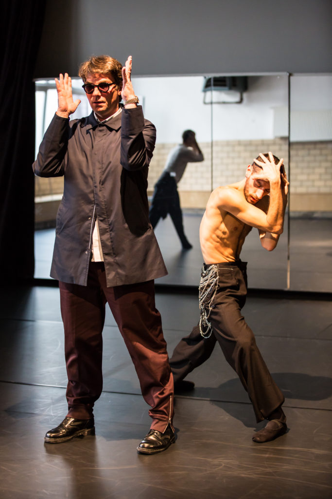 Marco Goecke, in street clothes and sunglasses, stands facing straight ahead, elbows raised so his palms frame his face. Dancer Luca Pannacci is slightly behind him in a deep fourth lunge, hands splayed over the top of his head as his elbows squeeze toward center.