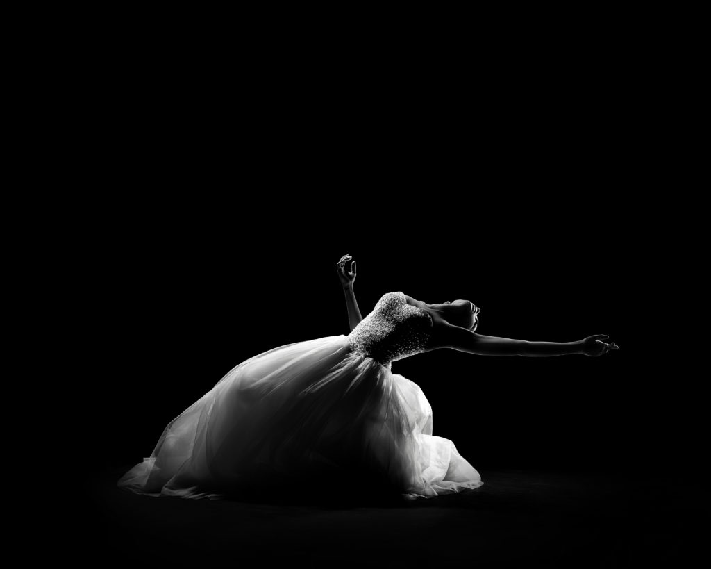 In a black and white image, a dancer in a white, flowing gown sinks to the floor in a shadowy space, an arm reaching plaintively behind her and to the side as she arches back.