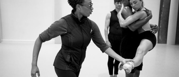In a black and white image, Amy Hall Garner is in profile, one hand adjusting the extended foot of a dancer being lifted. She wears an athletic vest over simple rehearsal gear and glasses.
