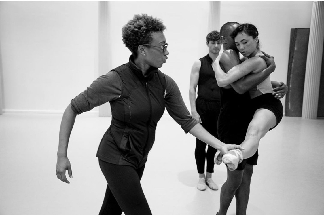 In a black and white image, Amy Hall Garner is in profile, one hand adjusting the extended foot of a dancer being lifted. She wears an athletic vest over simple rehearsal gear and glasses.