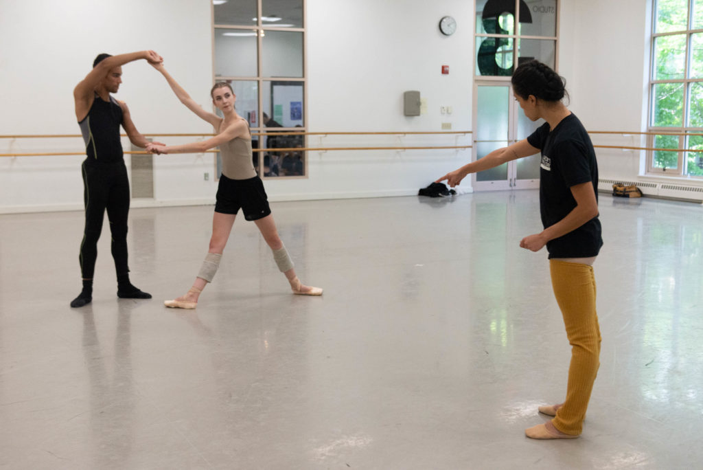 Lia Cirio stands in profile to the camera in an airy ballet studio, head tipped thoughtfully as she points towards a couple working out a partnering move. One of the dancers looks to Cirio, while the other watches his partner, holding her hands aloft.