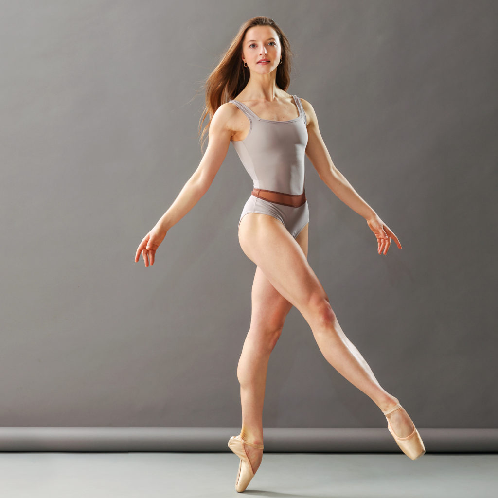 Indiana Woodward stands on pointe, low develope front and crossed, arms down and out loosely to the sides, her hair flowing down her back, looking at the camera
