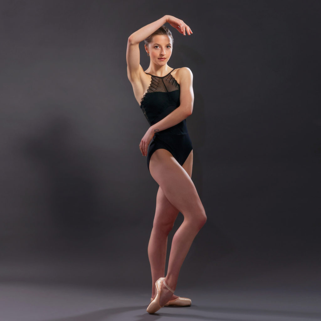 Indiana Woodward crosses her arms across her waist and above her head, front foot beveled, in a black leotard and pointe shoes on a grey background