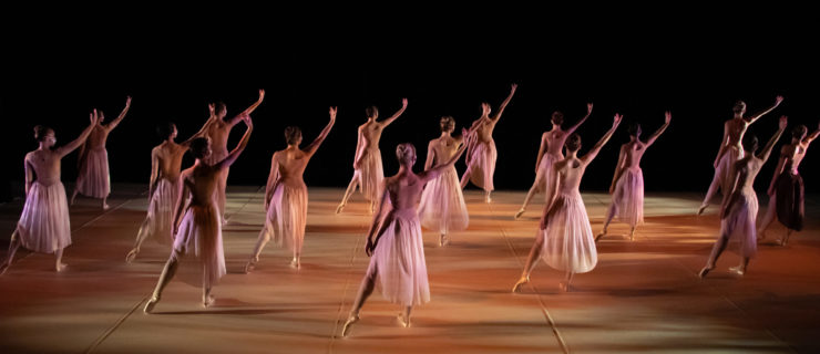 Female dancers in pale, flowing dresses and pointe shoes fill a simply lit stage. They face upstage, right arms reaching up on a diagonal, left legs extended in a parallel tendu side.