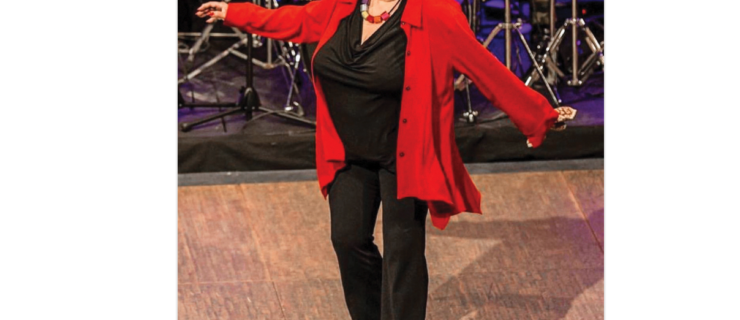 A mature Black woman with short, gay curly hair tap dances in front of a band. She is wearing white tap shoes, a black top and pants and a red, flowing cardigan.