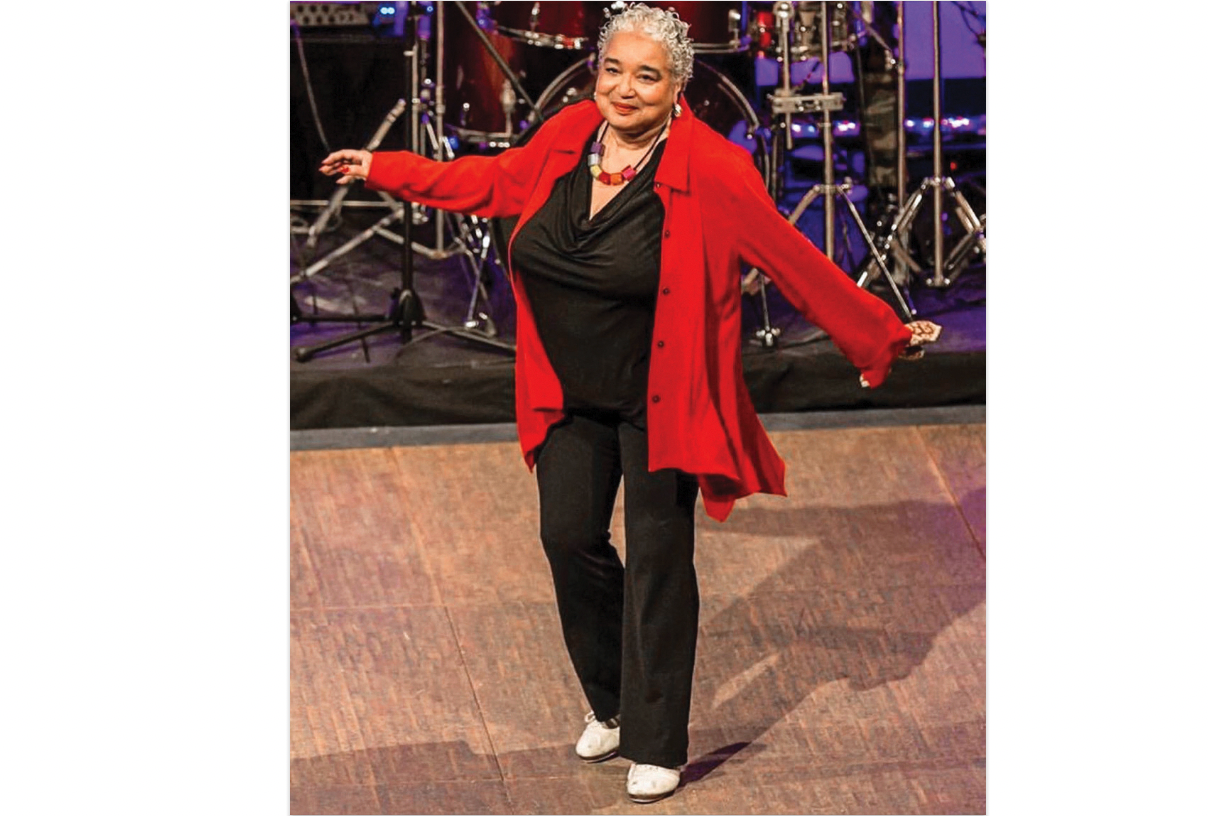 A mature Black woman with short, gay curly hair tap dances in front of a band. She is wearing white tap shoes, a black top and pants and a red, flowing cardigan.