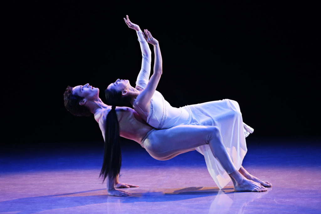 A male and female dancer in white pose together onstage in blue light. They face upward, the female dancer balanced across the male dancer's torso and thighs as he presses his hips up from the ground.