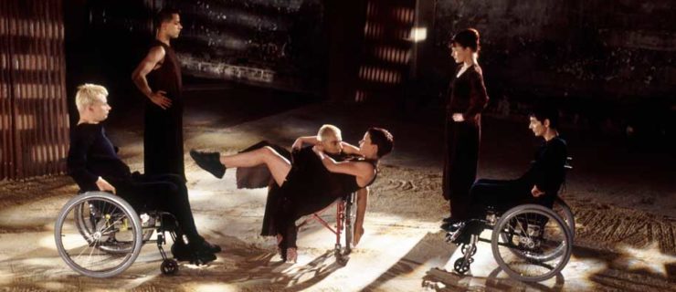 A film still shows six dancers in black in a sand-strewn interior space. To the left and right are two pairs of dancers facing in, those on the outside seated in wheelchairs and closer to center standing. They watch as a dancer lies almost horizontal to the floor, appearing to levitate from the other's chair.
