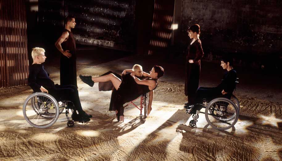 A film still shows six dancers in black in a sand-strewn interior space. To the left and right are two pairs of dancers facing in, those on the outside seated in wheelchairs and closer to center standing. They watch as a dancer lies almost horizontal to the floor, appearing to levitate from the other's chair.