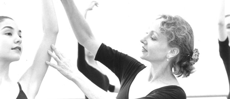 In a black and white image, Suki Schorer is seen in profile, both arms elegantly raised as she adjusts a student's arms in high fifth.
