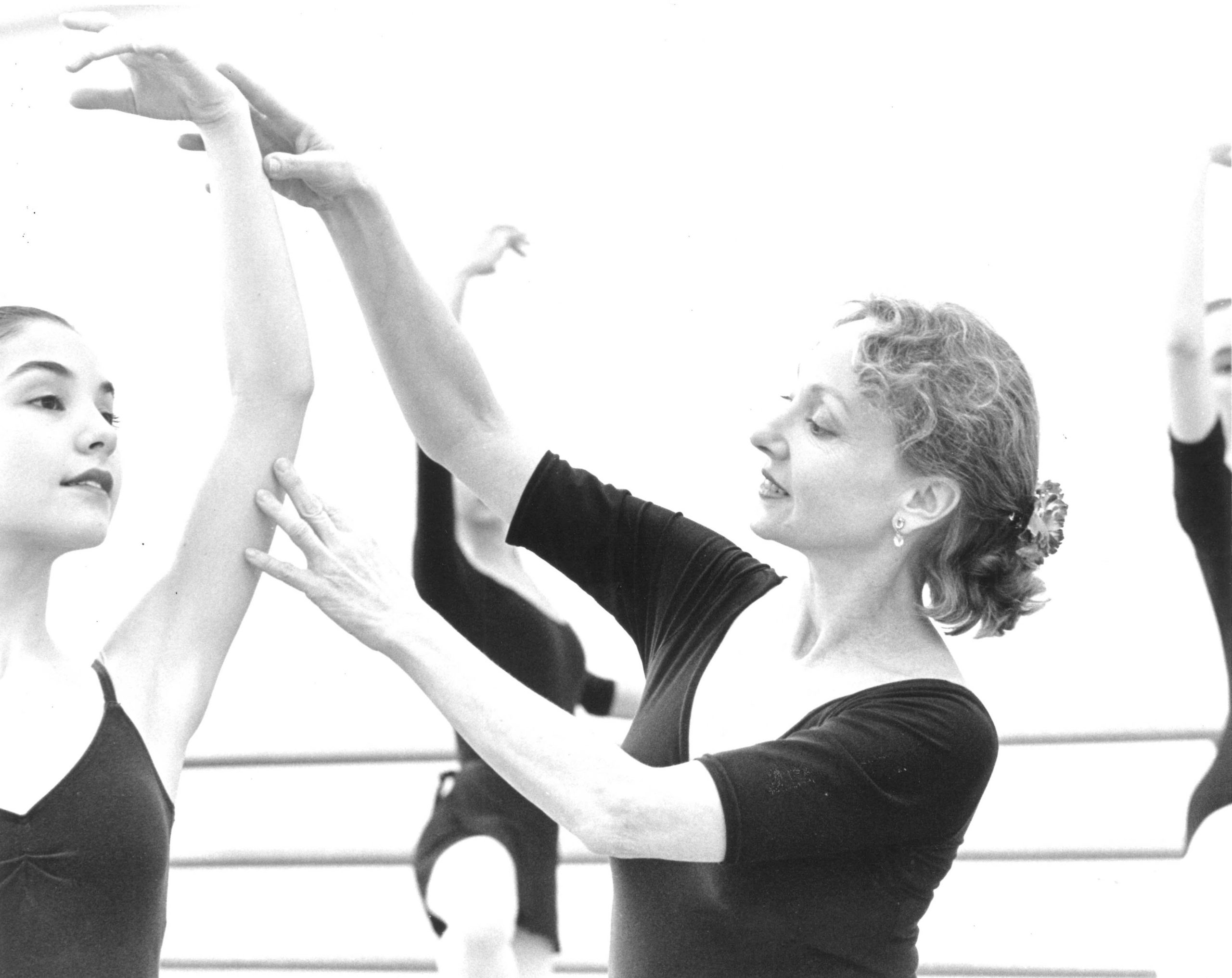 In a black and white image, Suki Schorer is seen in profile, both arms elegantly raised as she adjusts a student's arms in high fifth.