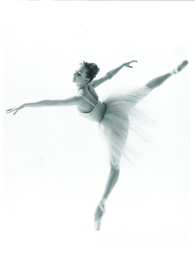 In a black and white image, a young Suki Schorer is in fourth arabesque en pointe, her torso tipped further forward in an almost French style.