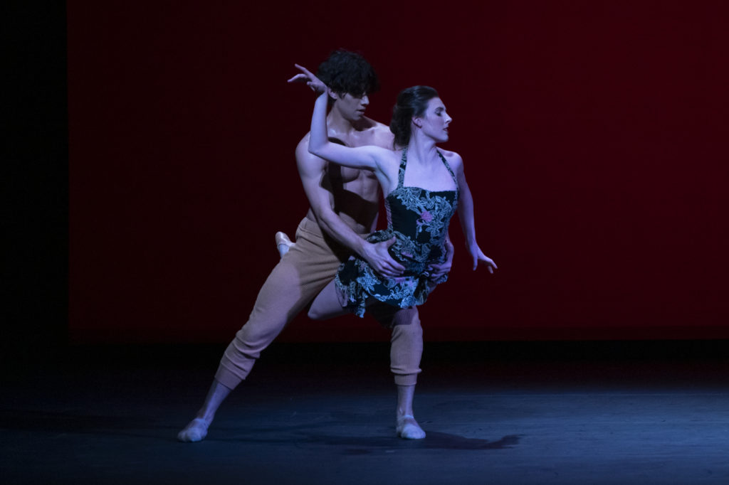 Roman Mejia stands in a low side lunge, balancing Tiler Peck over one knee with his hands holding her hips. Her legs bend around his supporting leg while she arches back, arms forming right angles as she looks cooly away.
