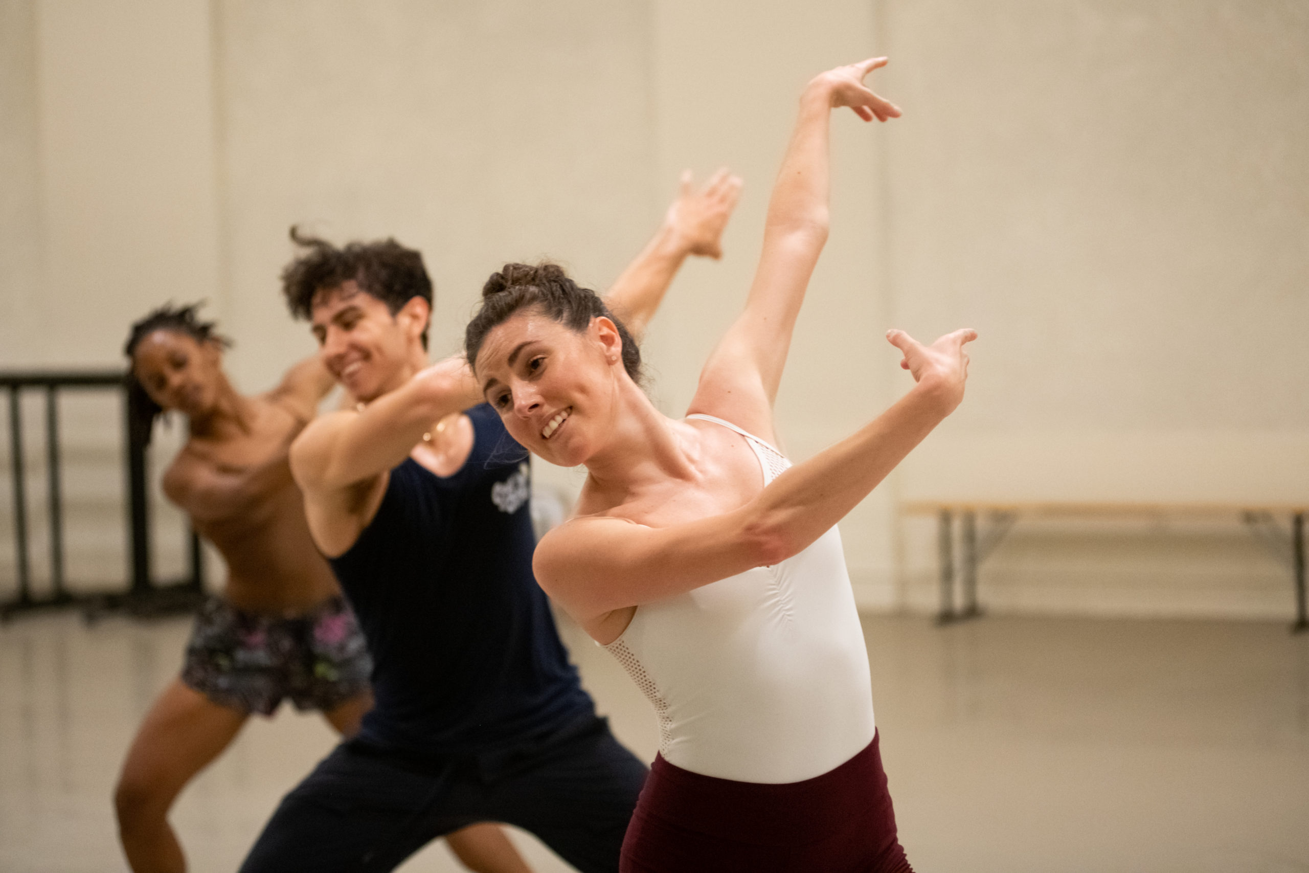 Shown from the hips up, Tiler Peck smiles as her arms swing in one direction as she leans in the other. Two dancers in a line behind her move through the same position, legs in a wide second position plié.