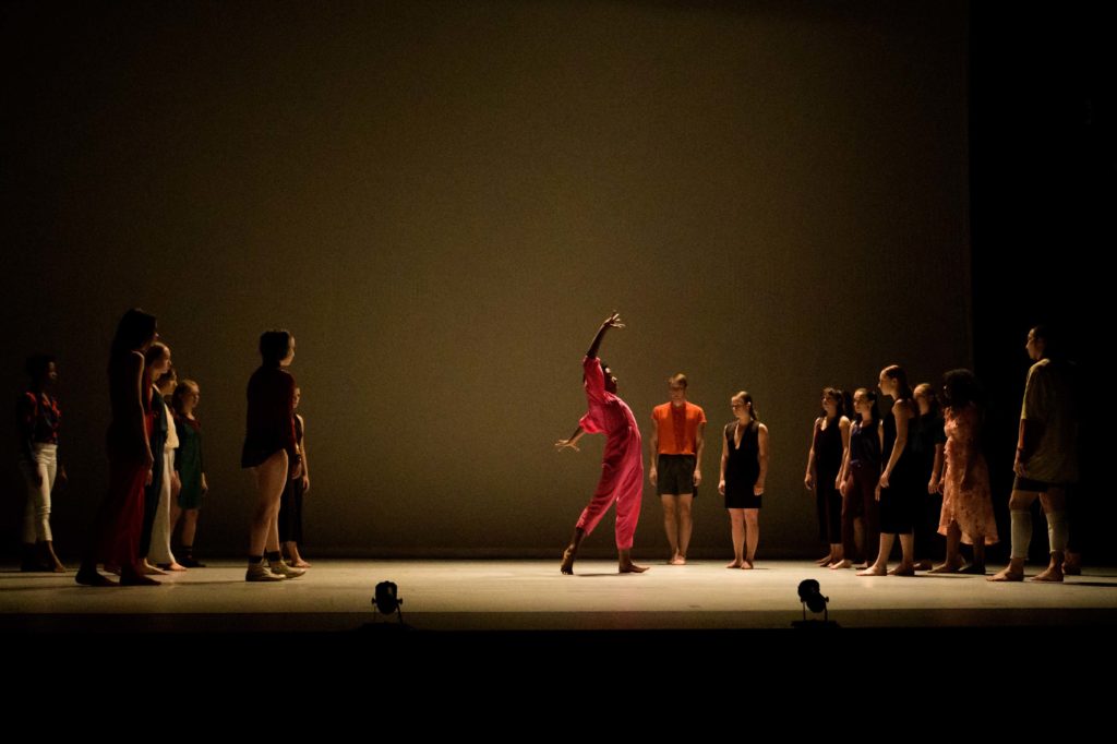 On a dimly lit stage, a dancer in bright red splays their ribs arms curving overhead and behind, leaning into their beveled back foot. They are surrounded by more than a dozen other dancers, all facing or walking slowly towards the dancer in red.