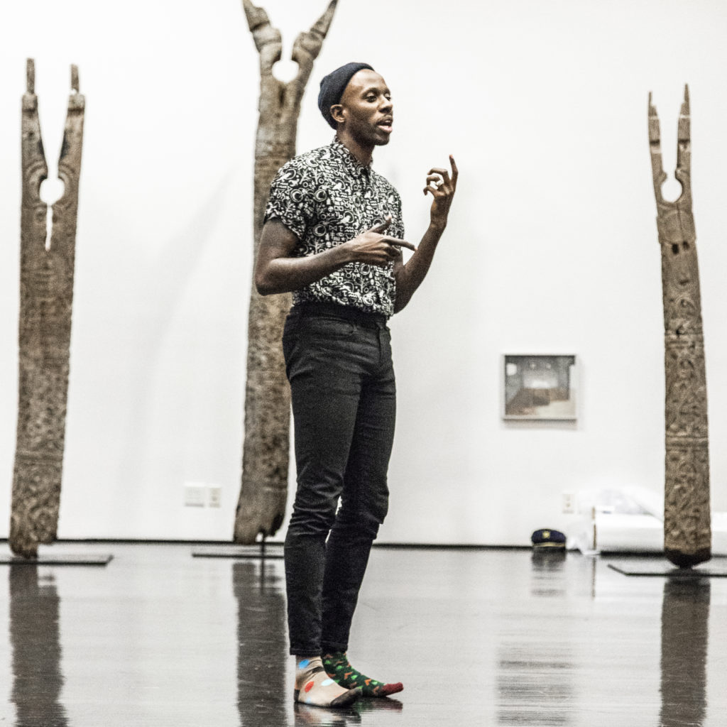 Raja Feather Kelly stands wearing colorful, mismatched socks on a shiny floor as he speaks in the middle of an art gallery, gesturing with his hands.