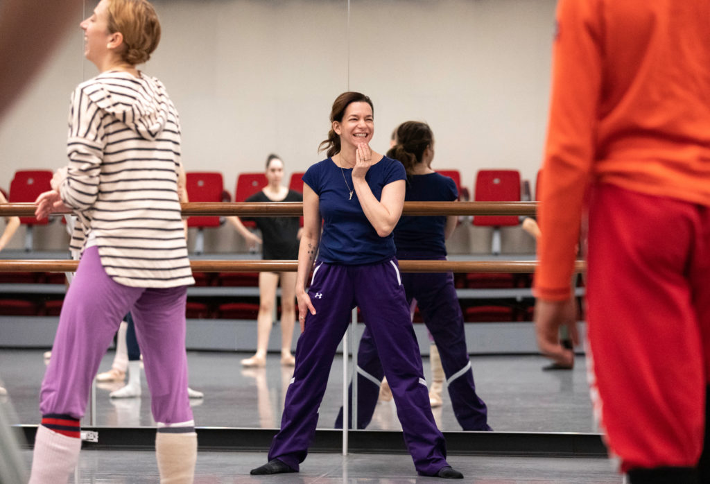 Pam Tanowitz stands in a wide second position at the front of a studio, smiling as she brings one hand to her chin. Dancers wearing layers are blurry in the foreground and in the mirror behind her.