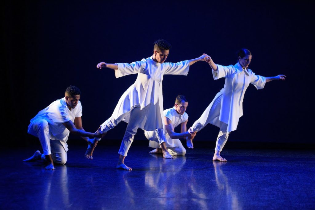 Four dancers in white pause on a blue-lit stage. Two balance with their downstage leg extended low behind them, leaning forward with their arms extended side, holding hands. Two other dancers kneel behind them, holding the standing dancers' ankles to provide a counterbalance.