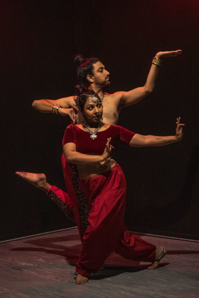 Amit Patel and Ishika Seth pose against a dark backdrop, both wearing red costumes and a combination of silver and gold jewelry. Seth looks over her left shoulder, extending her left arm with her palm upraised, her right hand matching the mudra. Patel balances on one leg directly behind her, face turned in profile towards his upraised right arm, elbow bent and palm to the ceiling.