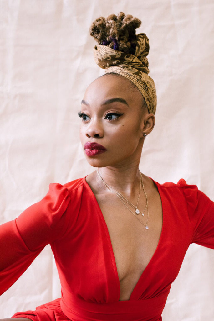 A portrait of Camille A. Brown. She gazes intently over one shoulder, not acknowledging the camera. Her red lipstick is a few shades darker than her long-sleeved blouse. Her braids are wrapped in a gold patterned head wrap so they sit piled at the back of her head.