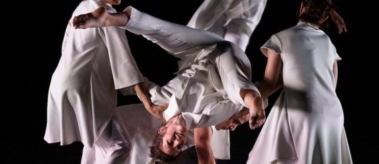 An upside-down dancer smiles brightly, legs in a controlled splay. The dancer balances on the back of another who is on all fours, while another on either side supports their shoulders. All wear different variations of all white outfits; the stage is dark behind them.