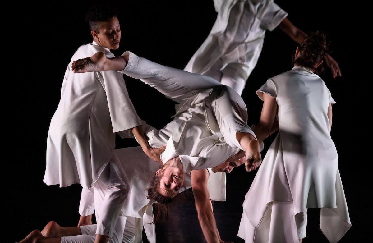 An upside-down dancer smiles brightly, legs in a controlled splay. The dancer balances on the back of another who is on all fours, while another on either side supports their shoulders. All wear different variations of all white outfits; the stage is dark behind them.