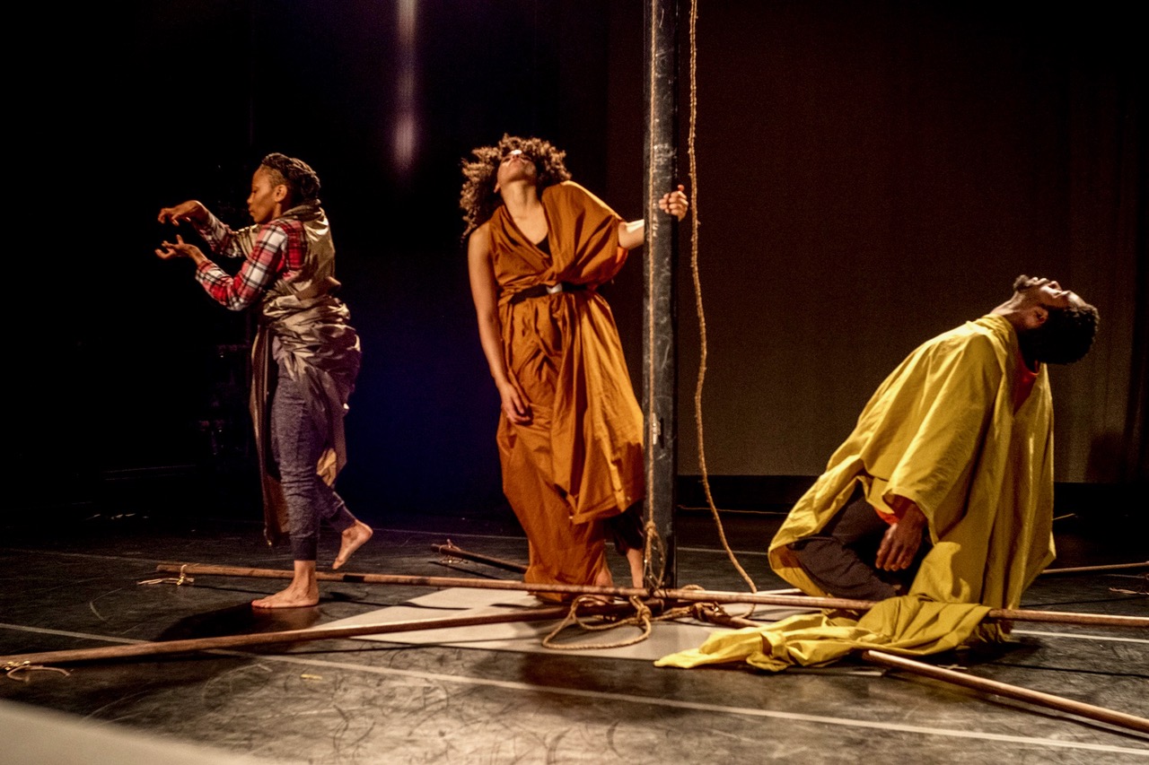 Three Black dancers are arrayed around a tall wooden pole resembling a ship's mast. On the floor around it are long, straight sticks and lengths of rope. A dancer robed in yellow hinges to the ground, a second in orange tips her head back as she grips the mast, and a third stares at a cradle formed by her empty hands, one foot lifted slightly off the ground.
