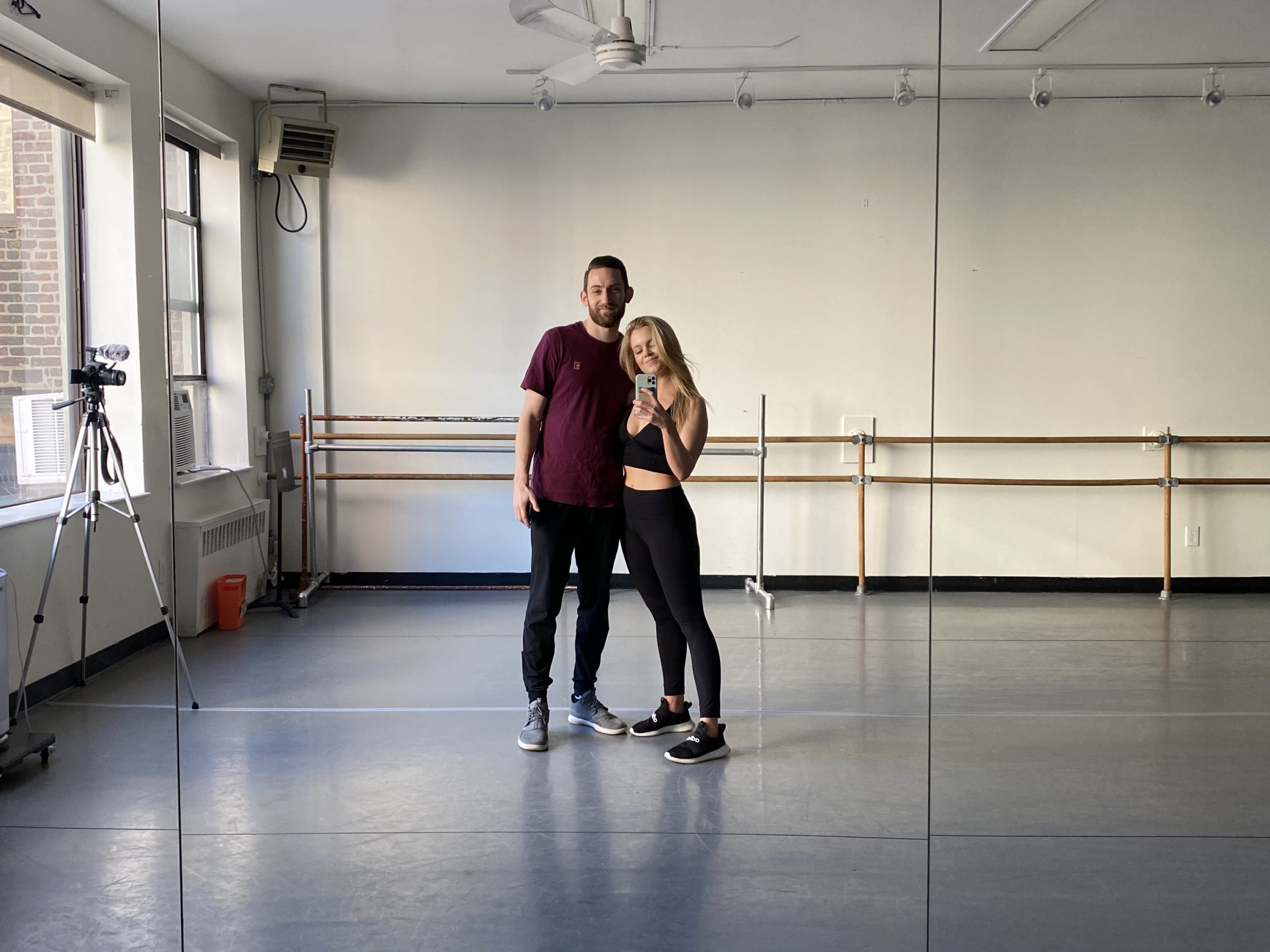 Haley Hilton wears black leggings and tank top, and her husband Coleman Clyde wears a loose maroon short-sleeved T-shirt and dark pants. They have an arm around each other and are taking a selfie in a dance studio, with ballet barres in the background.