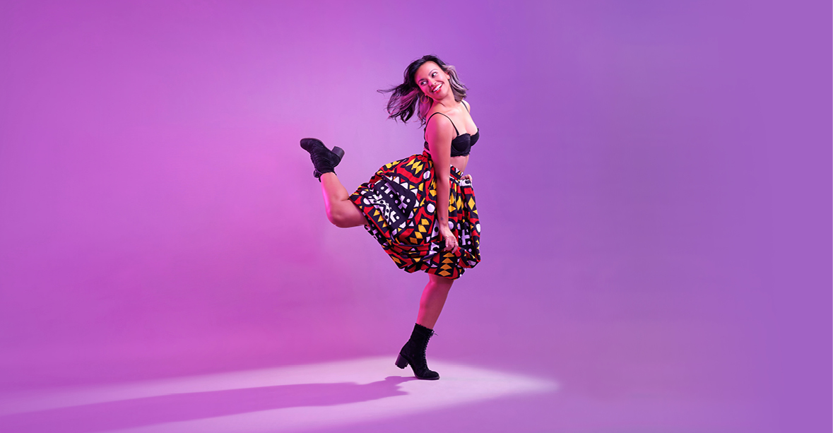 Ellenore Scott swings her back leg into a parallel attitude, her supporting leg in forced arch. She smiles over her shoulder at her back foot, while her downstage arm presses down against the colorfully patterned, knee-length skirt. She wears black lace-up heeled boots and a black bralette. Her black and silver hair waves down to her shoulders and flips behind her.