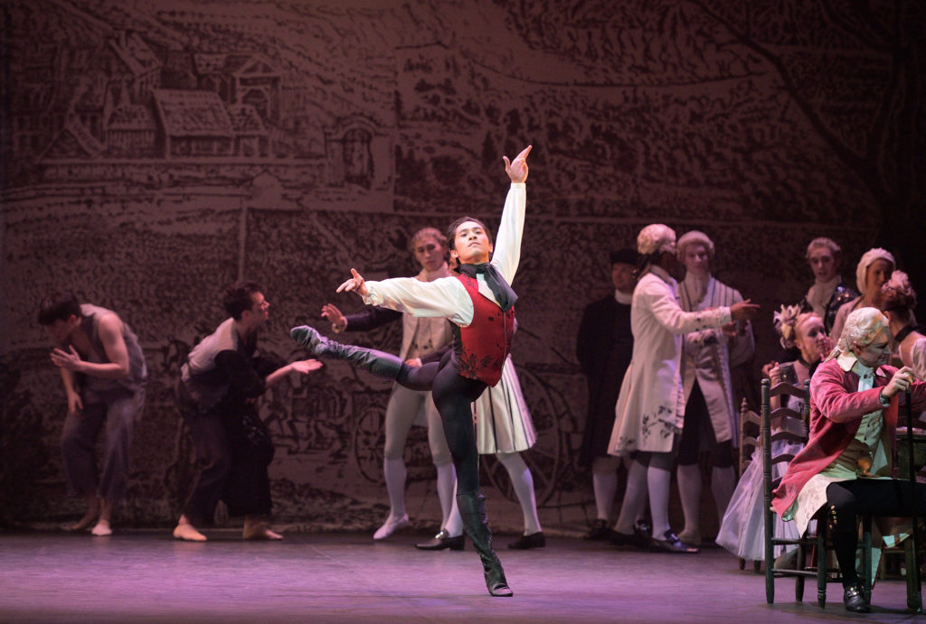 Jeffrey Cirio balances in croisé attitude back, arms in a presentational fourth. His hair is slicked back and he is costumed in black tights, ballet boots, and an ornate red vest and black cravat over a flowy white shirt. Dancers in 18th century period-wear mill in the background.