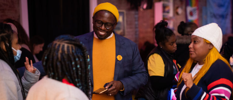 Reggie Harris smiles as he speaks with a cluster of women. He wears a yellow beanie that matches his sweater and a flower on the lapel of his navy blue suit jacket.