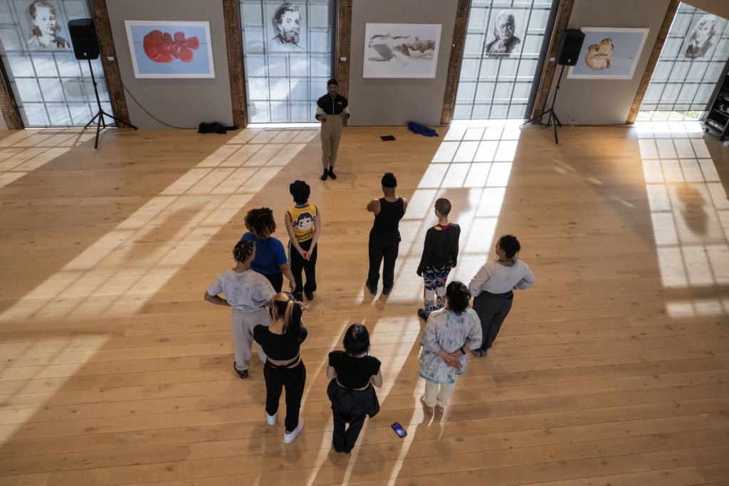 Omari Wiles addresses a circle of nine dancers, all in comfortable practice clothes. They are shown from above in a sunny studio space with wood floors and modern art on the walls.