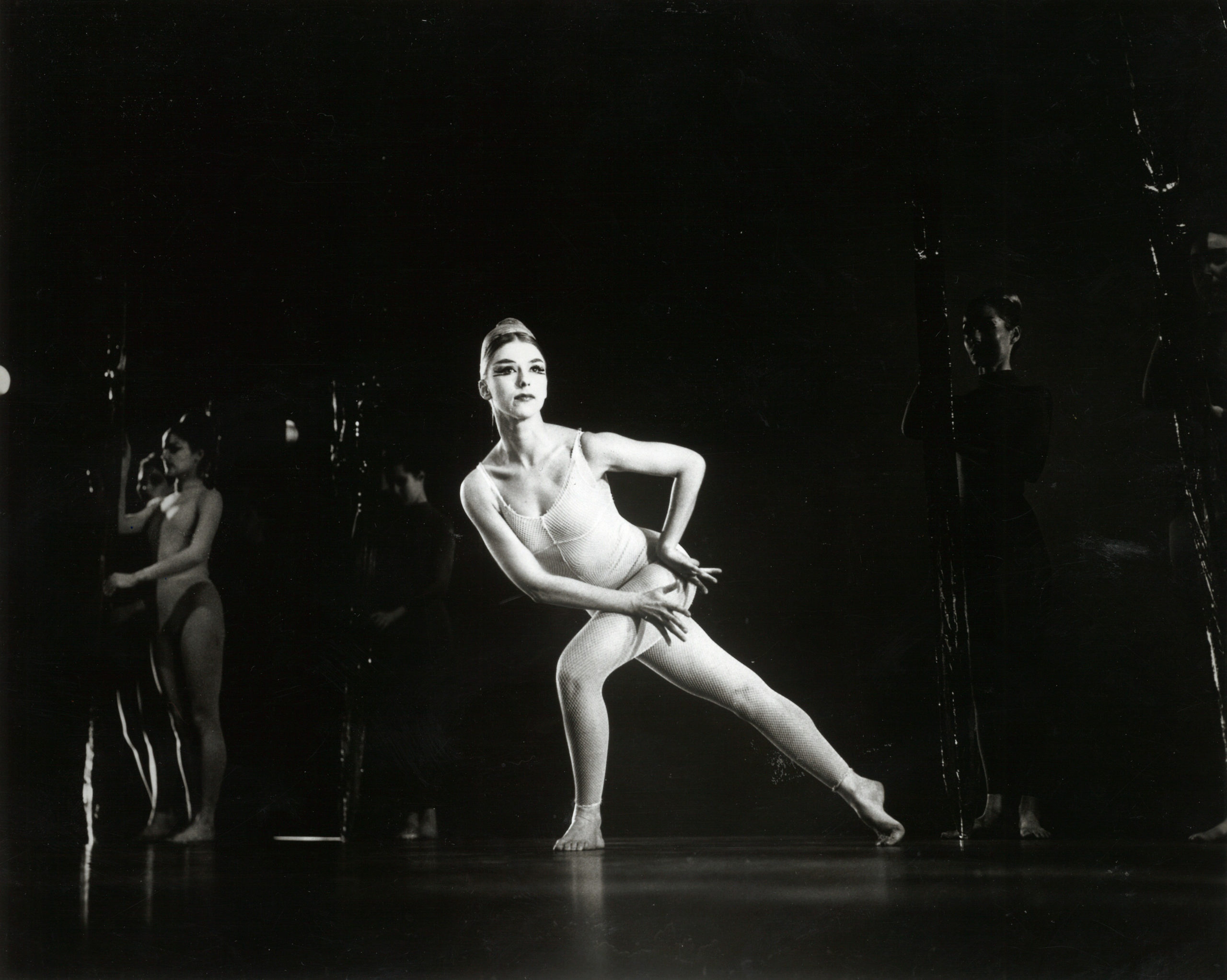 In a black and white archival image, Phyllis Lamhut is onstage in a pale unitard. She lunges forward, twisting so her torso forms a diagonal with her back leg and she can peer over her front shoulder. Her hands form a box at her downstage hip. Her blonde hair is neatly pulled back from her face.