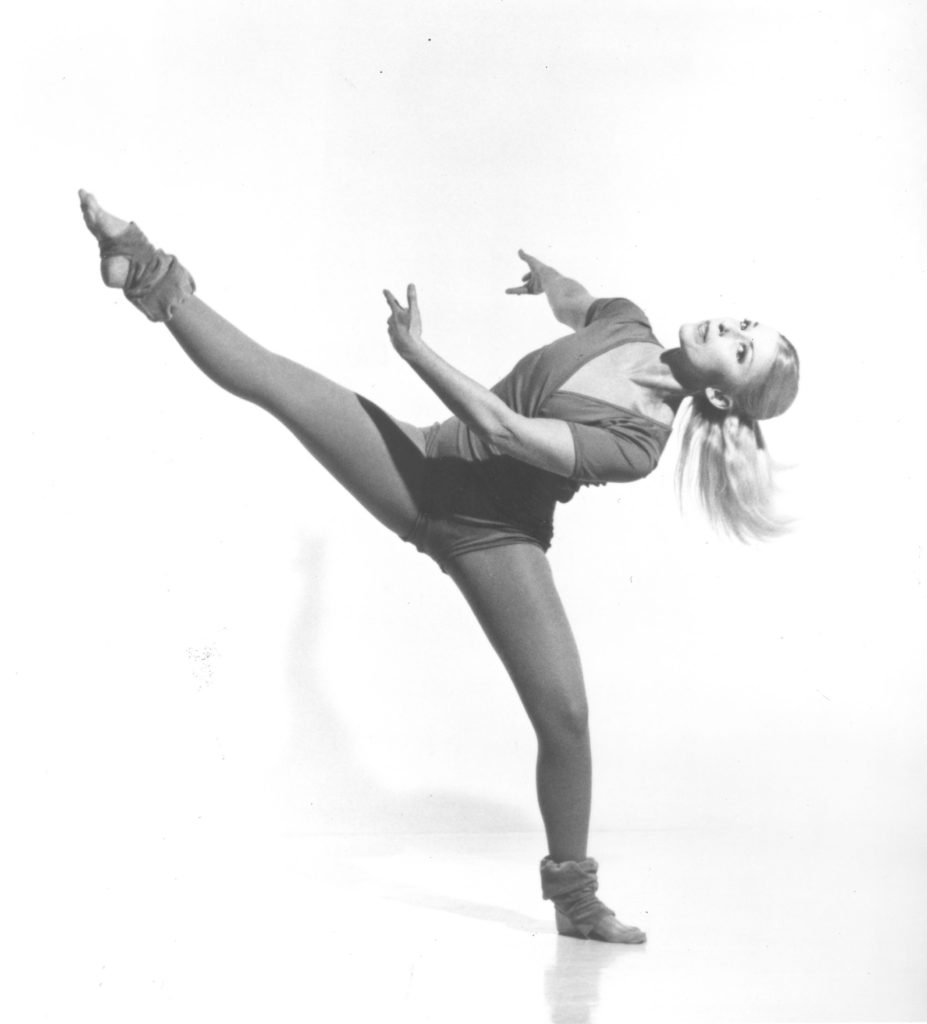 In a black and white archival image, Phyllis Lamhut is caught in an off-kilter extension. Her standing leg is in plié, working leg raised to the side while her upper body sways away from it. Her arms are gently curved, ponytail swinging, and she directs a calm, knowing look towards the camera.