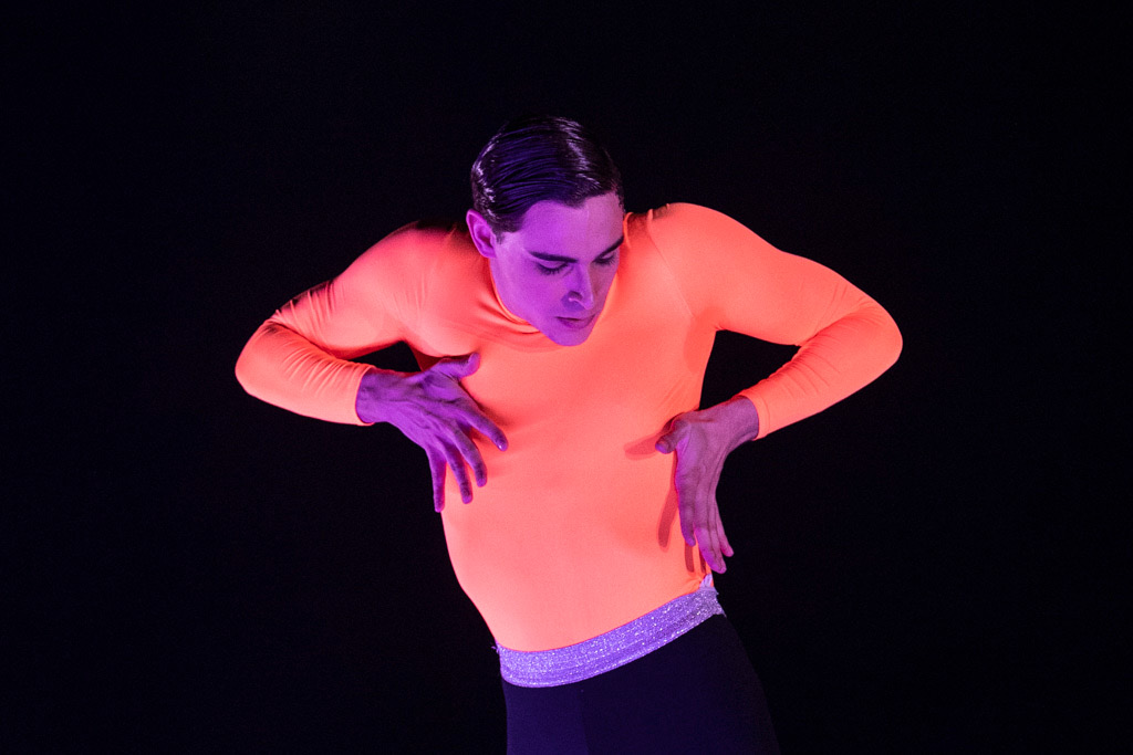 A dancer on a dark stage presses their palms against their ribcage, head jutting down towards them as their torso pulls away. Their skin is lit fuchsia in the stage lights, brightly contrasting with the close-fitting peach shirt they wear.