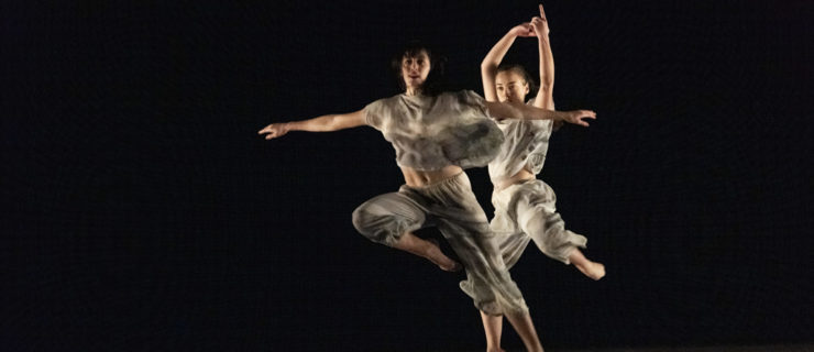 On a dark stage, two dancers in gauzy grey shirts and trousers are caught mid-air. The more downstage of the two extends her arms side as her right knee tucks up underneath her, left leg extended straight on a low diagonal. She partially blocks the dancer behind her, whose arms fly in a curve overhead as her knees bend slightly.