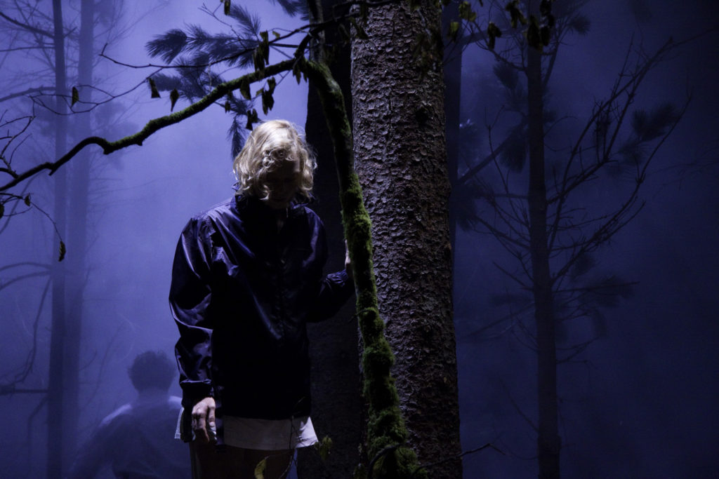 A blond person with their face in shadow stands with a hand resting on the trunk of a tree, head bowed. A mossy branch extends before them. In the smoky background, more trees and the head and shoulders of someone retreating.