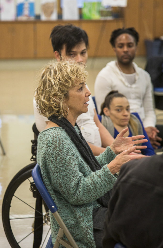 Heidi Latsky is shown in profile, gesturing with her hands as she speaks to a group seated in a circle with her.