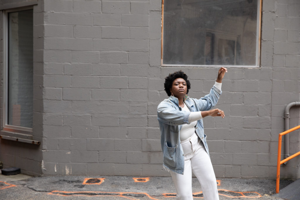 Ogemdi Ude looks at the camera, weight falling into her right leg as she lightly raises her bent arms to her left. She wears a light blue denim jacket over white jeans and a white shirt. Behind her, an industrial-seeming grey-painted wall and the beginning of an orange-railing to either steps or a ramp.
