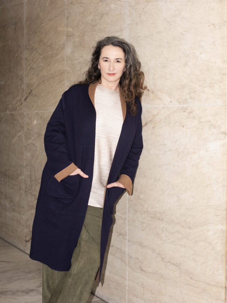 Adolphe Binder leans against a marble wall, silver and brown curls falling to her shoulders and hands in the pockets of her blue coat as she directs a small smile to the camera.