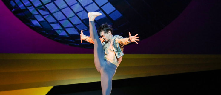 A male dancer battements his leg forward with a flexed foot, bottom leg in plié. He contracts his upper body slightly as the leg reaches 180 degrees, arms outstretched to his sides. In the background, an image of a massive blue disco ball.
