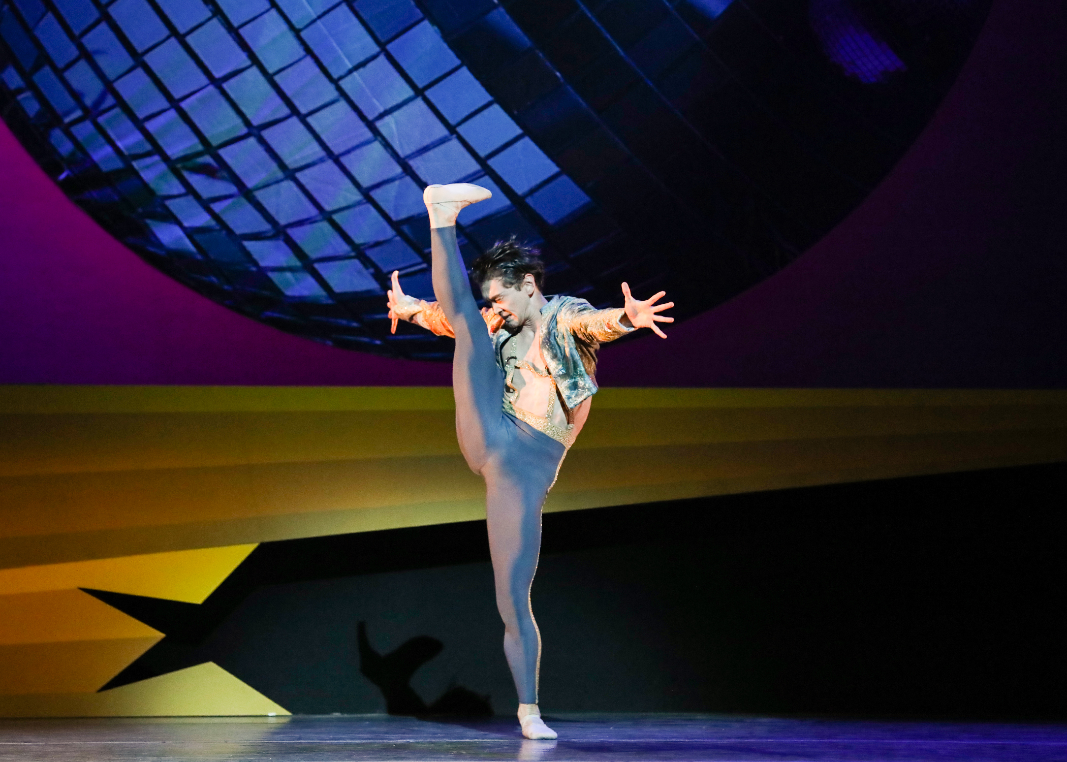A male dancer battements his leg forward with a flexed foot, bottom leg in plié. He contracts his upper body slightly as the leg reaches 180 degrees, arms outstretched to his sides. In the background, an image of a massive blue disco ball.