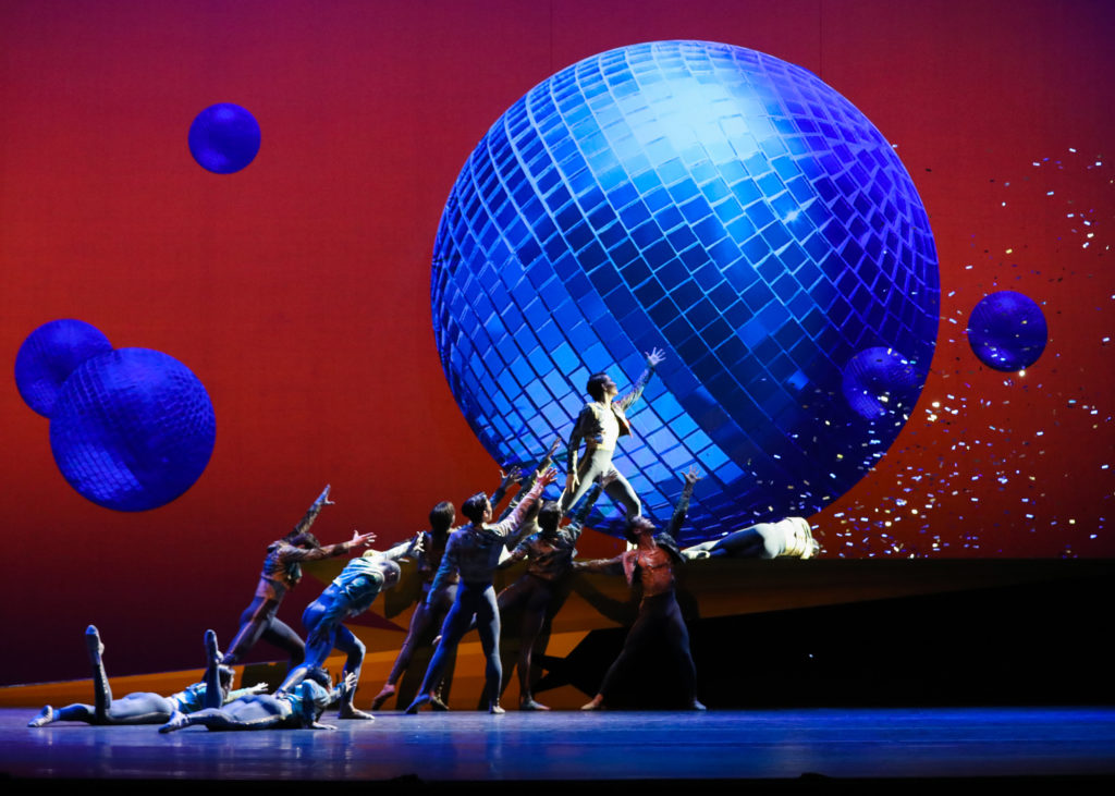 Ten male dancers strike poses at various levels, from prone on the floor to standing atop a raised platform, as they reach towards the upstage right corner of the stage with open hands. The backdrop is red with a large blue disco ball. Confetti flies from stage right.