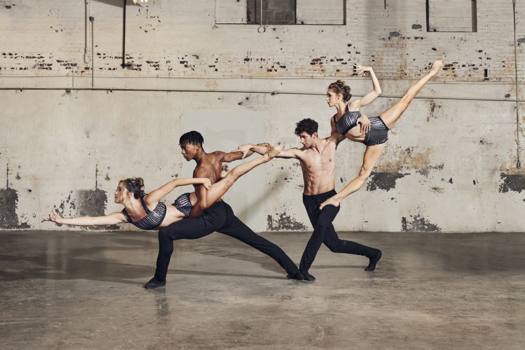 Four dancers form a chain in an industrial-seeming space. The women are lifted, reaching to the left side, while the men support them in deep lunges. The overall effect is that of an extended diagonal line.