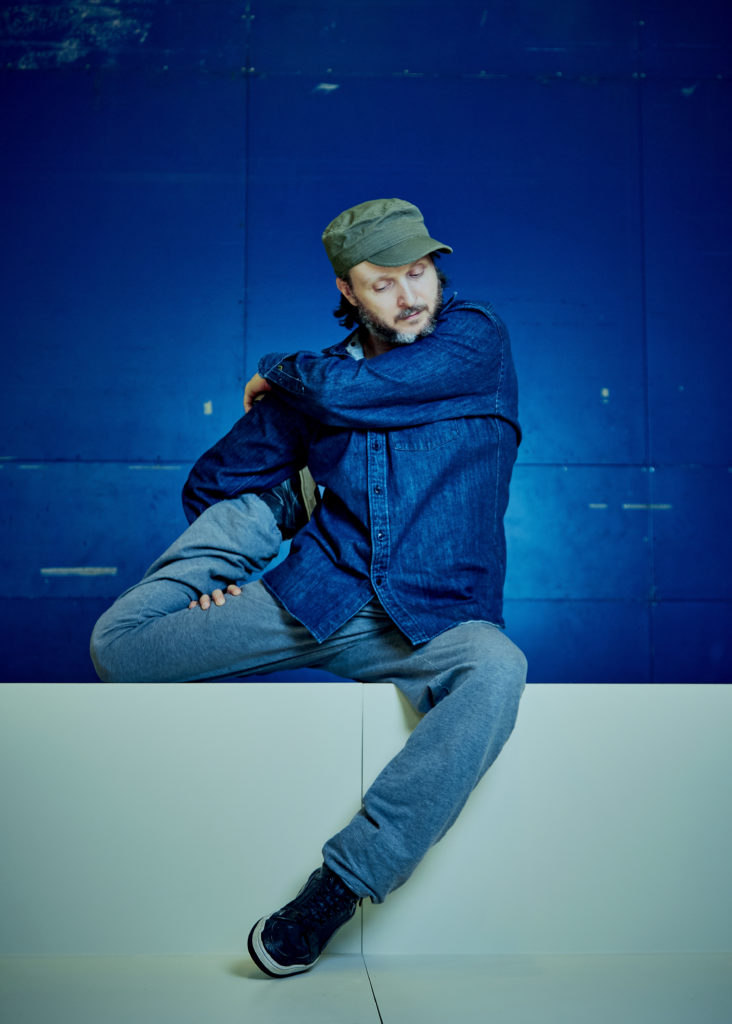 Sidi Larbi Cherkaoui sits on a ledge against a bright blue backdrop. One bent knee dangles off the ledge while his other leg bends so his heel touches his hip. His arms wind around the stretched leg, eyes downturned as his chin nearly rests on his crossed over arm. He wears a denim shirt, jeans, a green cap, and sneakers.